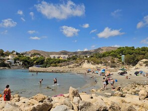 A beach with people swimming and sunbathing, a rocky shoreline, and buildings in the background.