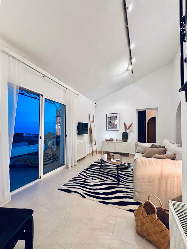 A modern living room with a sofa, a coffee table, and a zebra-patterned rug. There is a sliding glass door leading to a balcony with a view.
