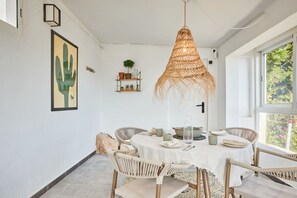 Un comedor con una mesa redonda, sillas de mimbre, una lámpara colgante y vistas a través de una ventana.