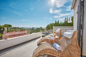 A rooftop terrace with wicker furniture, a small table with a bottle and glasses, and a view of the landscape.