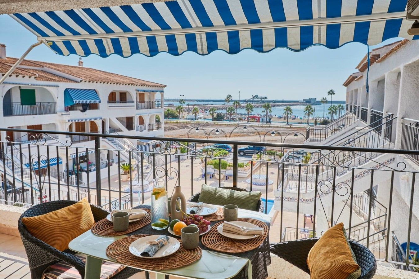 A balcony with a table set for two, overlooking a coastal area with buildings and palm trees.