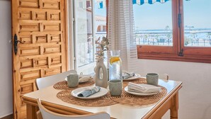 Une table à manger dressée avec des assiettes, des tasses et une carafe de thé glacé, offrant une vue sur la mer à travers de grandes fenêtres.