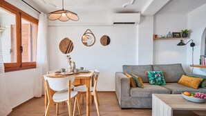 Un salon moderne avec un canapé gris, une table à manger en bois et des chaises. On y trouve une fenêtre avec des rideaux, un luminaire suspendu et une étagère murale agrémentée d’objets décoratifs.