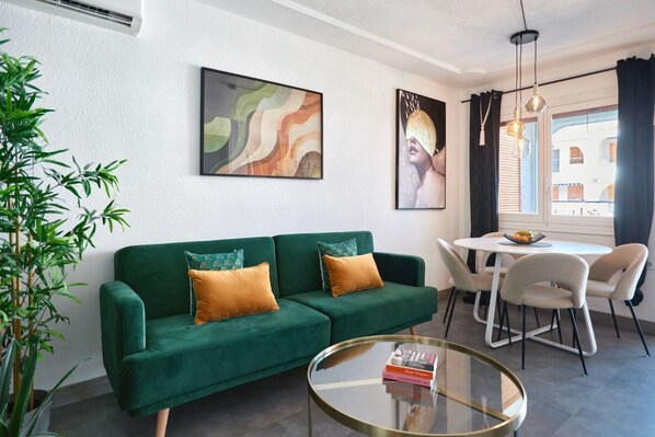 A green sofa with two cushions, a glass coffee table with a book, a dining table with chairs, and a large window with curtains.