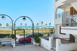 Une entrée fermée par un portail, avec une voiture rouge garée à l’intérieur, un balcon offrant une vue sur la plage et un ciel dégagé.