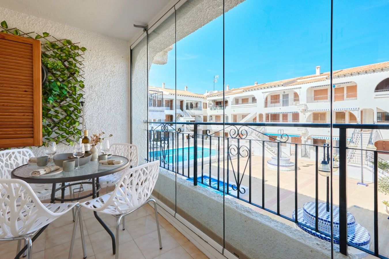 A balcony with a table, chairs, and a view of a pool and buildings.