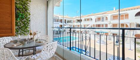 A balcony with a table, chairs, and a view of a pool and buildings.