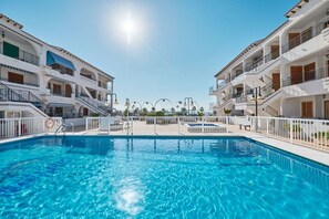 A clear blue swimming pool surrounded by white buildings with balconies and a clear sky.