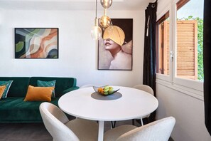 Une pièce avec un canapé vert, une table ronde entourée de chaises, un panier de fruits et des œuvres d’art accrochées aux murs.