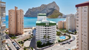 Ein Hochhaus-Hotel mit Pool, umgeben von einem Parkplatz und mit Blick auf das Meer und ein Gebirge.