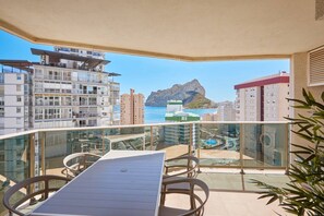 A balcony with a table and chairs, overlooking a coastal cityscape with buildings and a mountain.