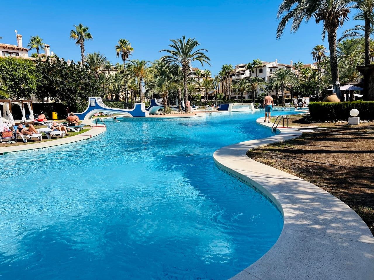 A large swimming pool with a curved edge, surrounded by palm trees and lounge chairs.