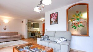 A living room with a sofa, a coffee table with magazines, a kitchen in the background, and a large floral artwork on the wall. - 1 bedroom gorgeous apartment in Masso (Masso)