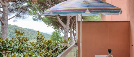 A balcony with a striped umbrella, a table set with plates and a bottle, and a view of a forested hillside.