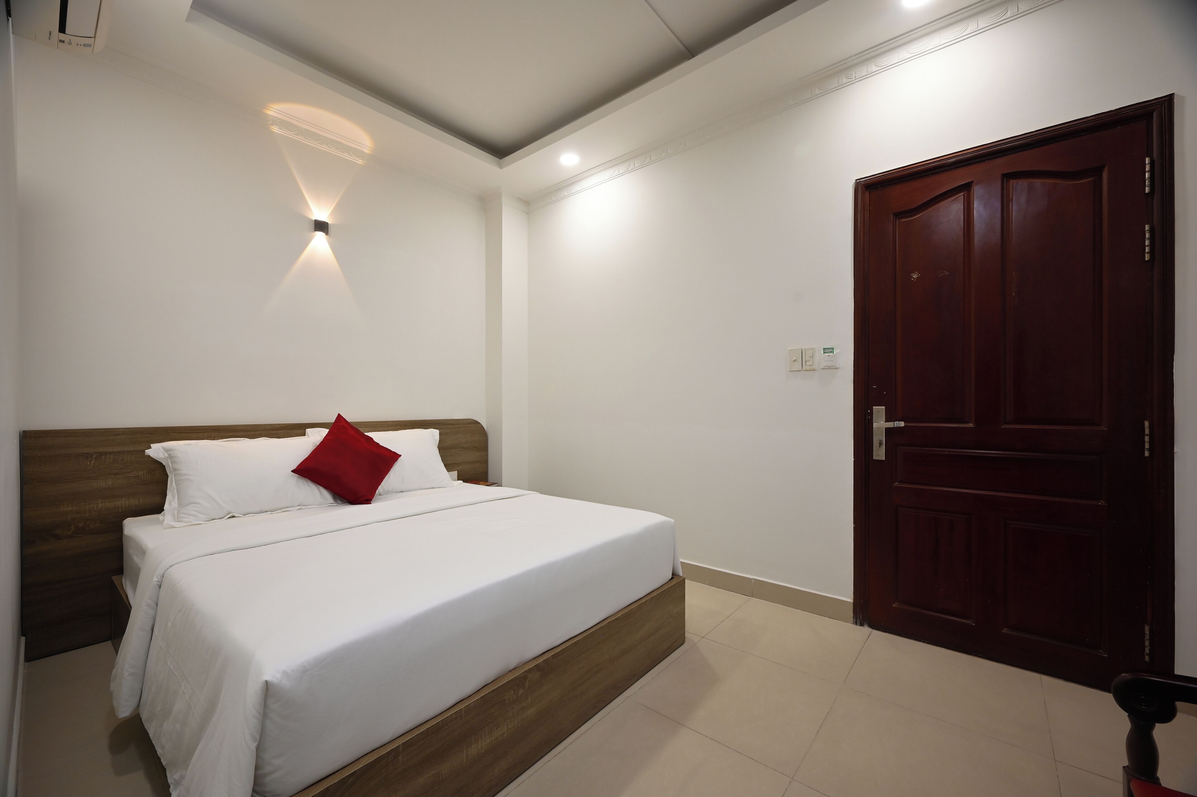 Een slaapkamer met een houten bed, wit beddengoed, een rood kussen, een wandlamp en een donkerhouten deur.