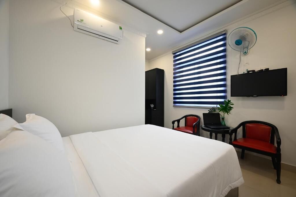 A hotel room with a bed, a TV mounted on the wall, a window with blinds, and a small table with a chair.