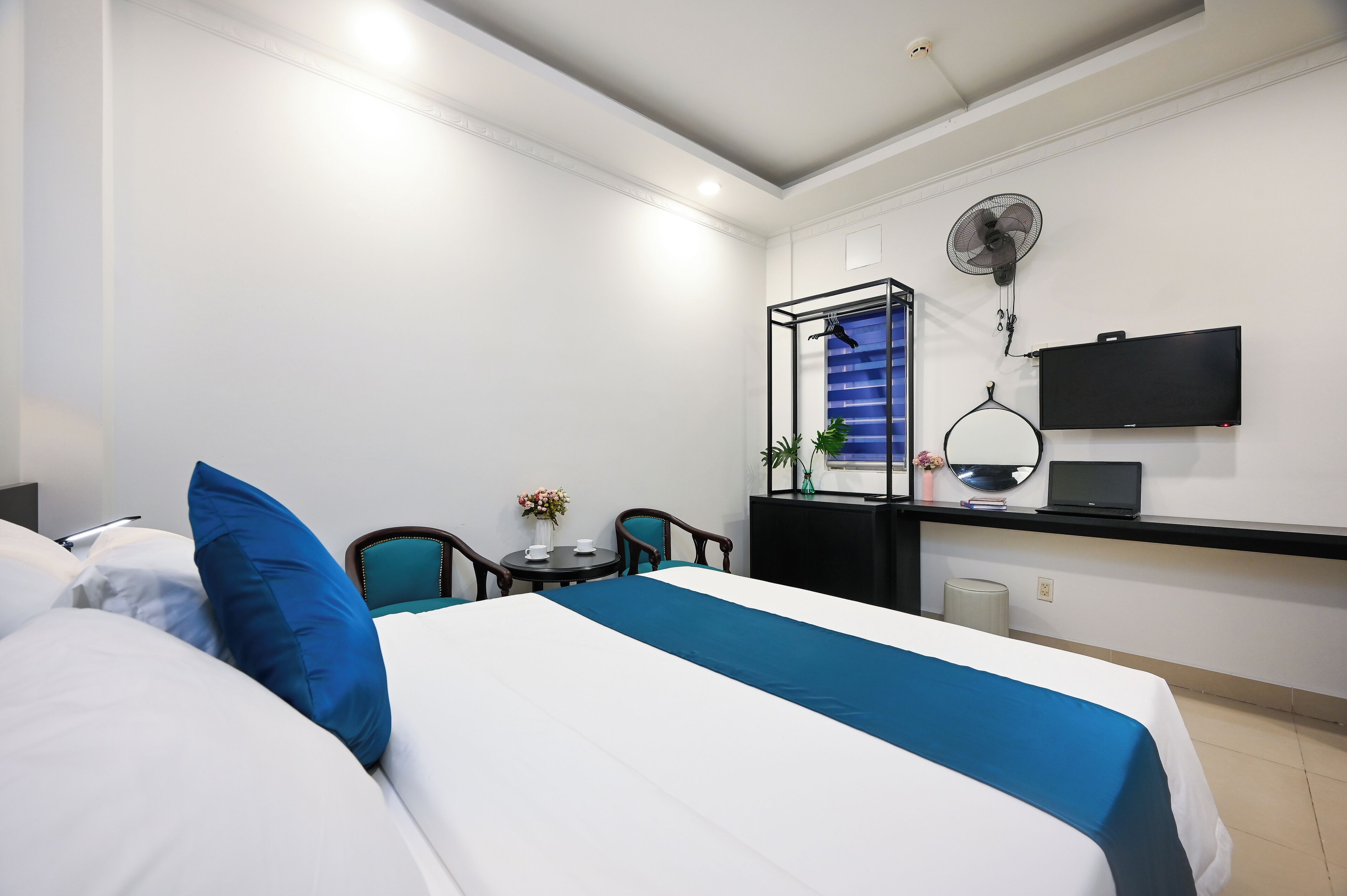 A hotel room with a bed, a desk with a laptop, a TV, a fan, and a small table with a vase.