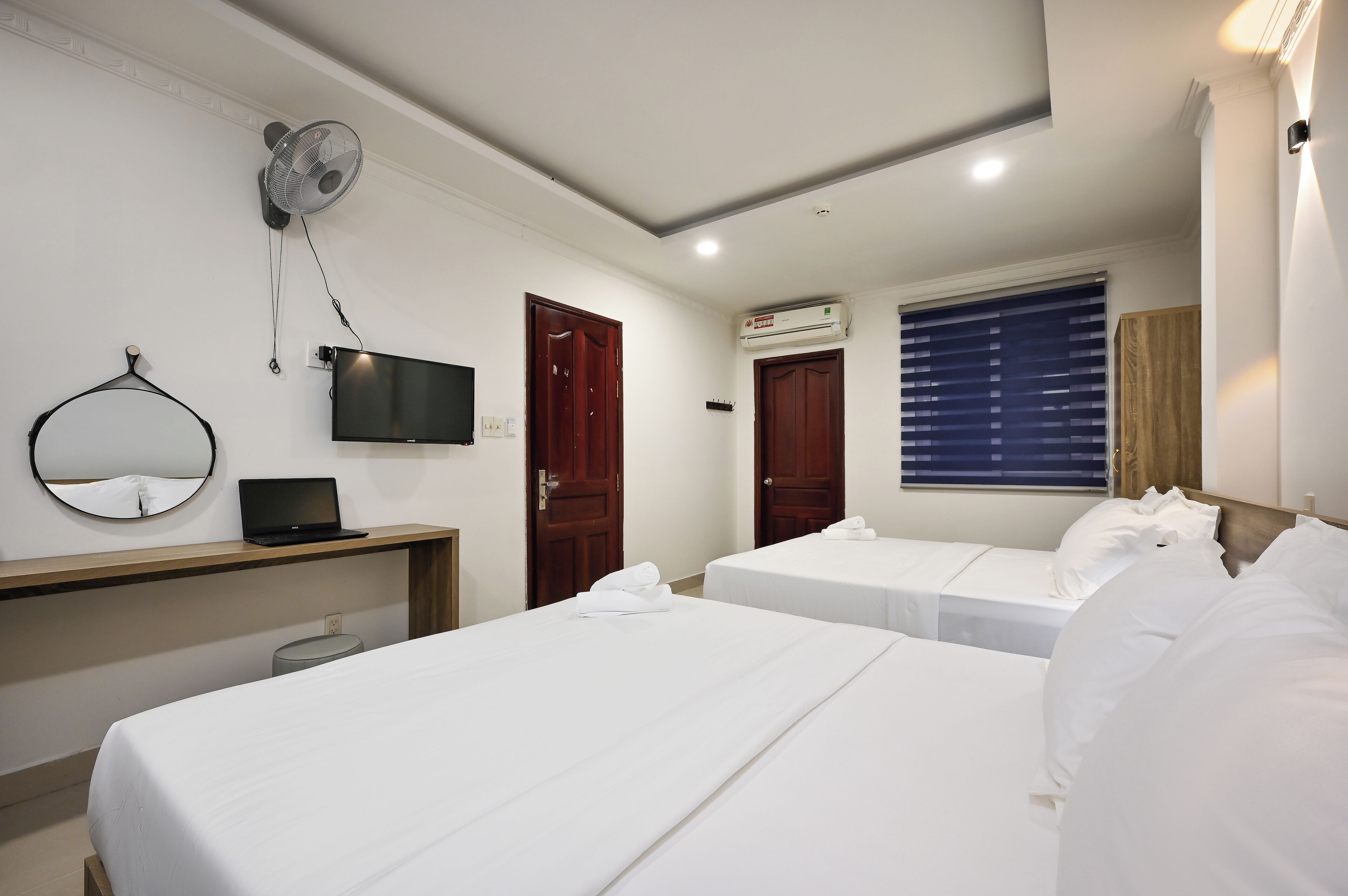 A hotel room with two beds, a desk with a laptop, a TV, a wall-mounted fan, and an air conditioning unit.