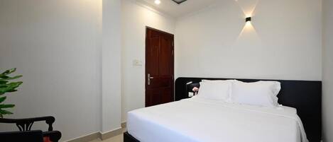 A hotel room with a bed, a desk with a laptop, a door, and a wall-mounted light fixture.