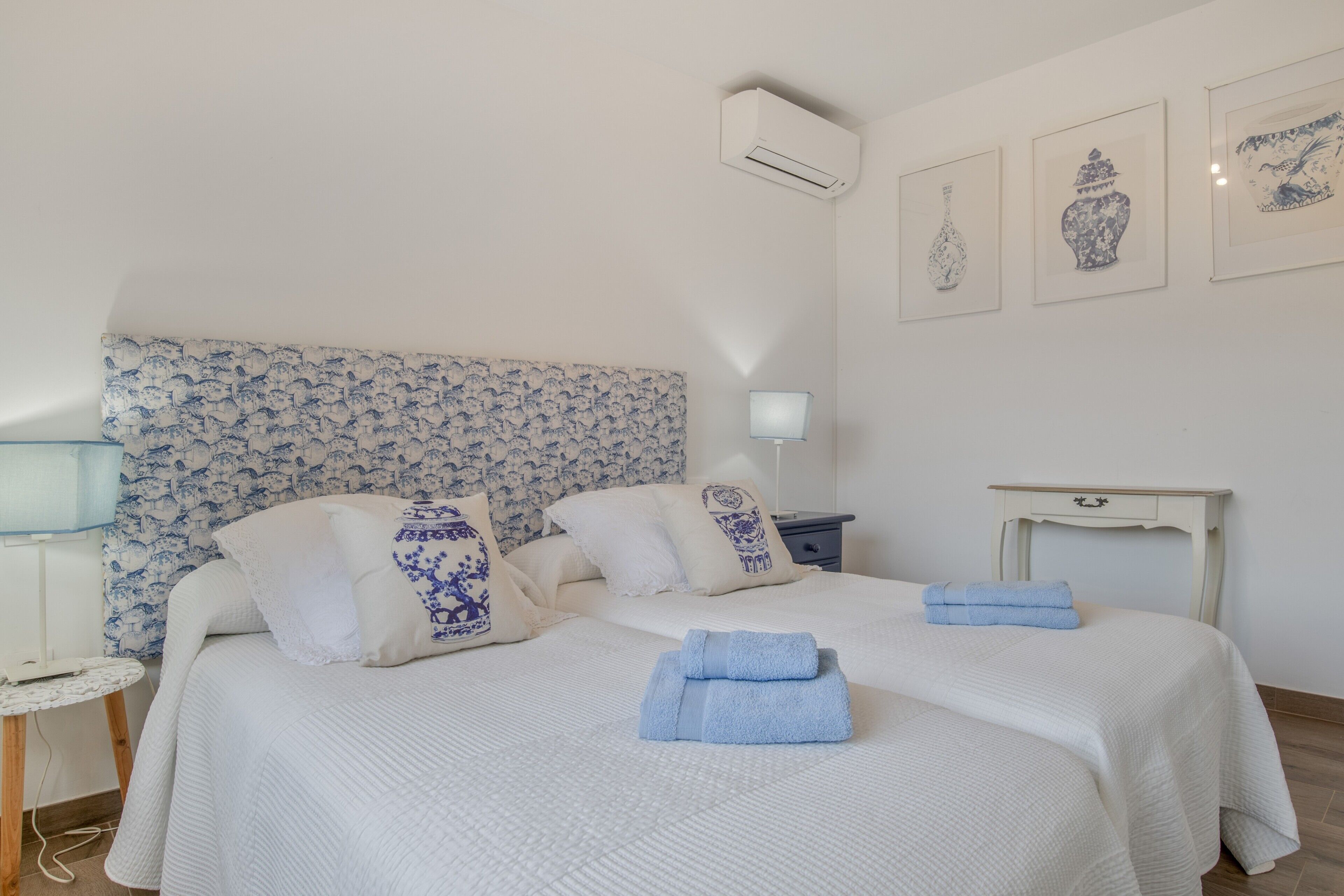 A neatly made bed with a floral headboard, a bedside table with a lamp, and wall-mounted artwork.