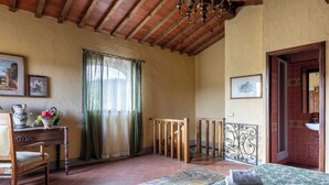 A room with a wooden bed, a desk with a chair, a window with curtains, and a bathroom visible through an open door. - La Casina Cottage by Geco Vacation Rentals (Bucine)