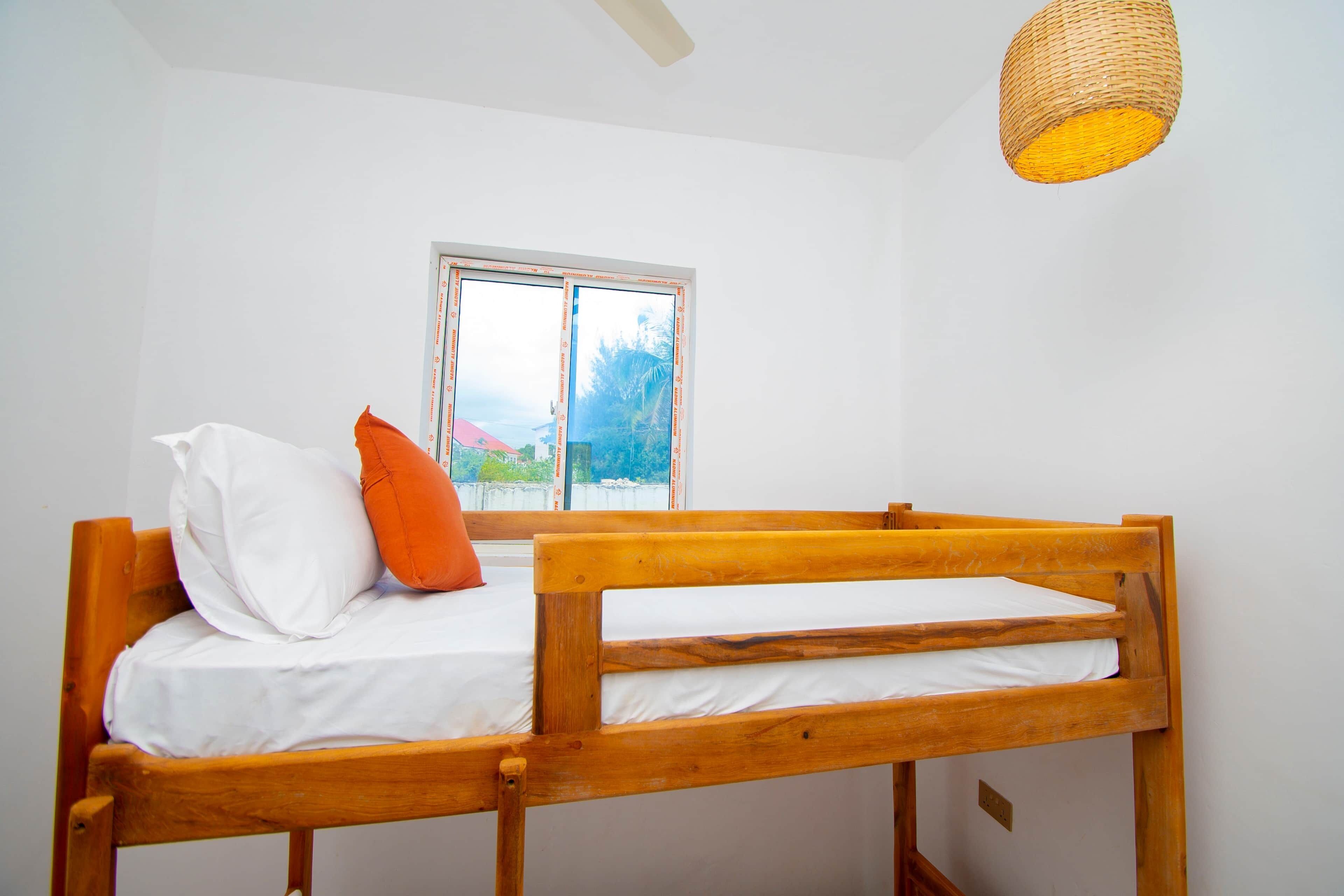 A single wooden bed with white bedding and an orange pillow in a room with white walls and a window.