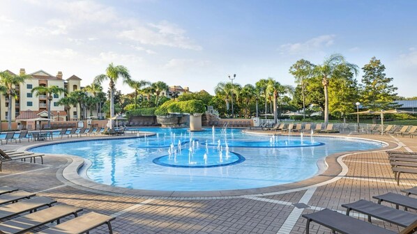 A swimming pool with multiple fountains, surrounded by lounge chairs and umbrellas, with palm trees and a building in the background.