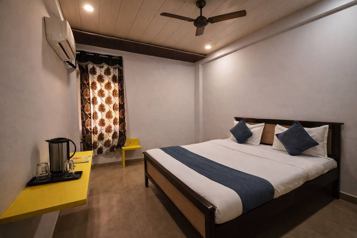 A hotel room with a bed, a desk with a coffee maker, a chair, a ceiling fan, and a window with patterned curtains.