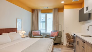 A hotel room with a bed, two armchairs, a desk, a chair, a sink, and a window.