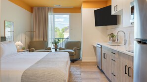 A hotel room with a bed, two armchairs, a small table with a plant, a kitchen area with a sink and a TV mounted above it. - Brand New Modern Stay In 12 South | The Gilmore (Nashville)