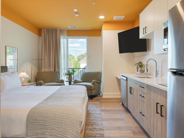 Une chambre d’hôtel avec un lit, deux fauteuils, une petite table avec une plante, un coin cuisine avec un évier et un téléviseur fixé au-dessus.
