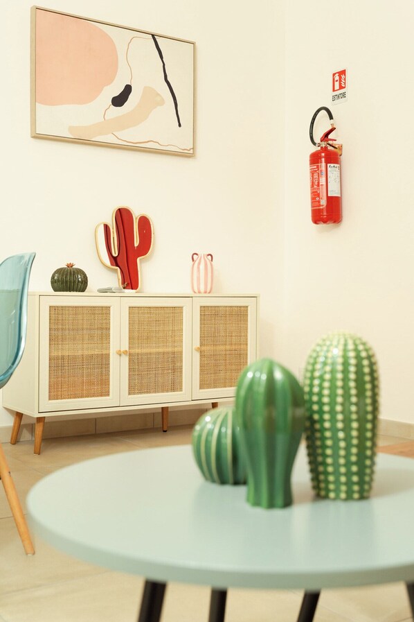 Une pièce avec une table ronde, un meuble blanc orné d’un motif de cactus, un extincteur et un tableau abstrait accroché au mur.