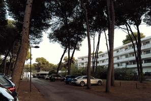Un espace de stationnement avec plusieurs voitures, de grands arbres et, au fond, un immeuble à plusieurs étages.