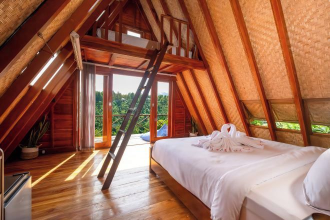 Una habitación con techos altos, una escalera de madera, una cama con sábanas blancas y vistas al exterior con vegetación.