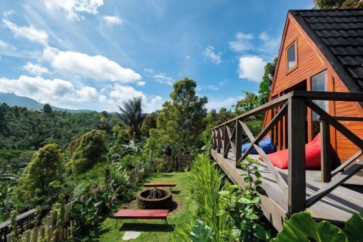 A wooden cabin with a deck, surrounded by lush greenery and a clear sky.