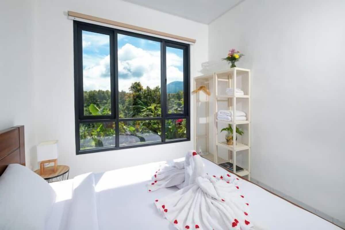 A hotel room with a bed, a window with a view of greenery, a white shelf with towels, and a small table with a lamp.