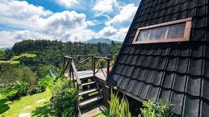 Um edifício com telhado preto e escadaria de madeira que leva a um deck, cercado por vegetação exuberante e montanhas ao fundo.