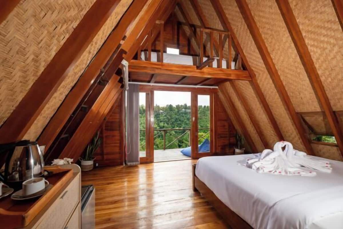 A cozy attic bedroom with a wooden ceiling, a bed with white linens, a small desk with a kettle, and a view of greenery outside.