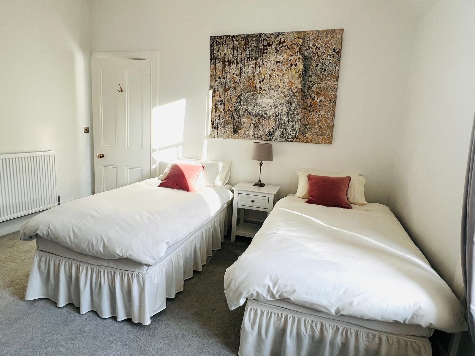 Une chambre avec deux lits, une porte blanche, un radiateur, une table de chevet avec une lampe et un grand tableau abstrait accroché au mur.