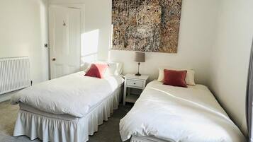 Une chambre avec deux lits, une porte blanche, un radiateur, une table de chevet avec une lampe et un grand tableau abstrait accroché au mur.