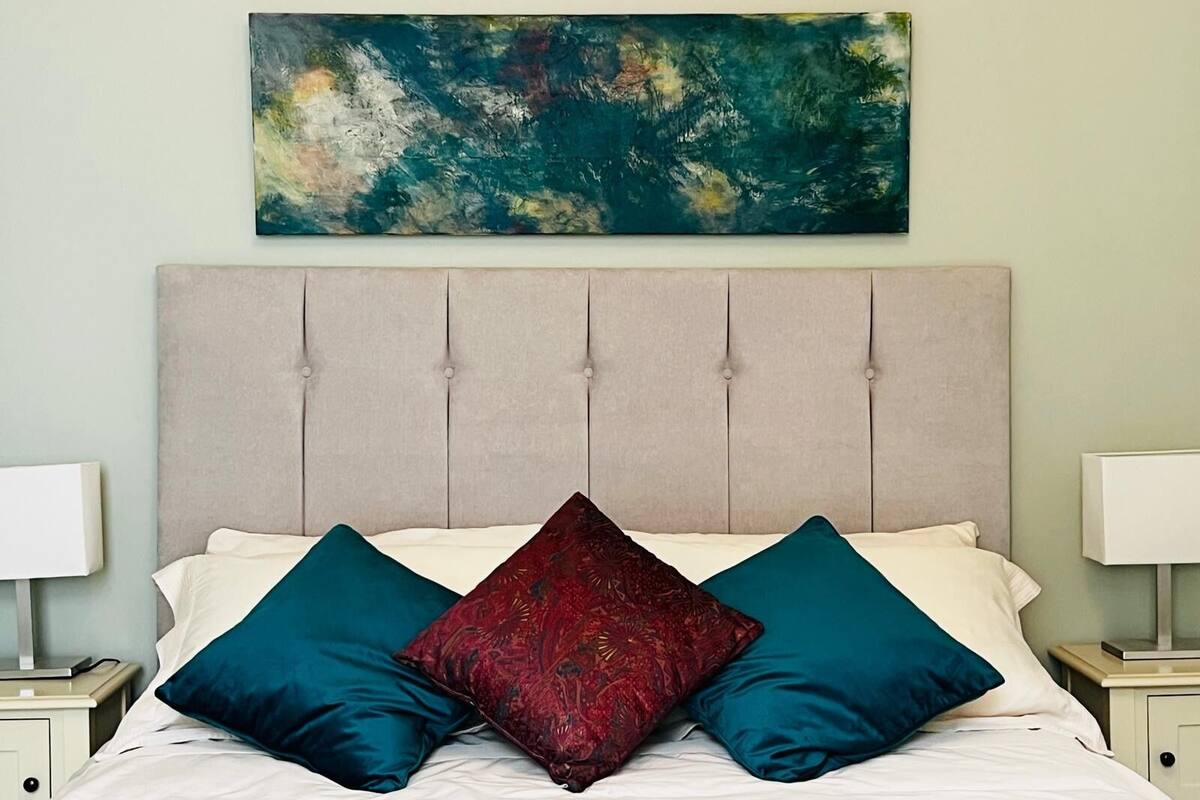 Una cama con cabecera capitonada, dos almohadas, una mesita de noche con lámpara y un cuadro en la pared.