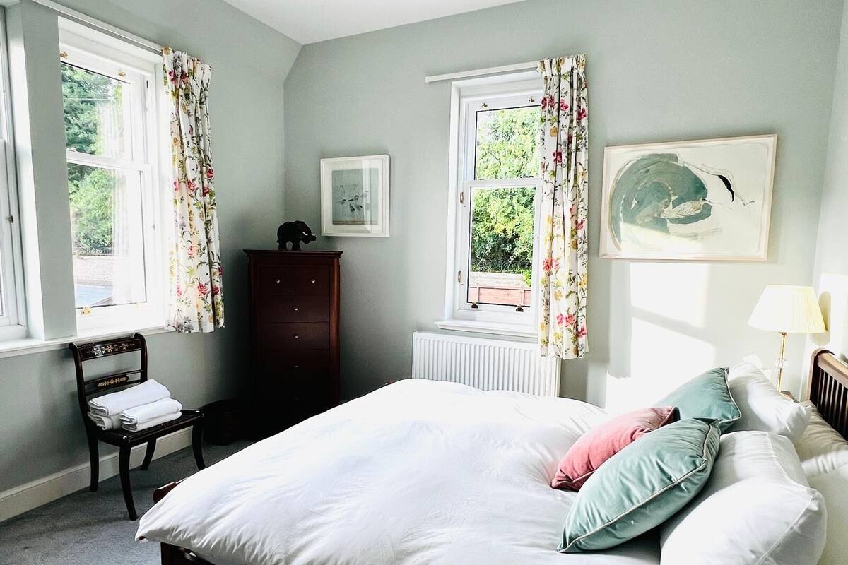 Un dormitorio con cama, una silla, cómoda, dos ventanas con cortinas, un radiador y un cuadro en la pared.