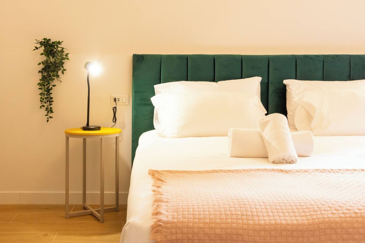 A neatly made bed with white linens and a green headboard, a small table with a lamp, and a potted plant on the wall.