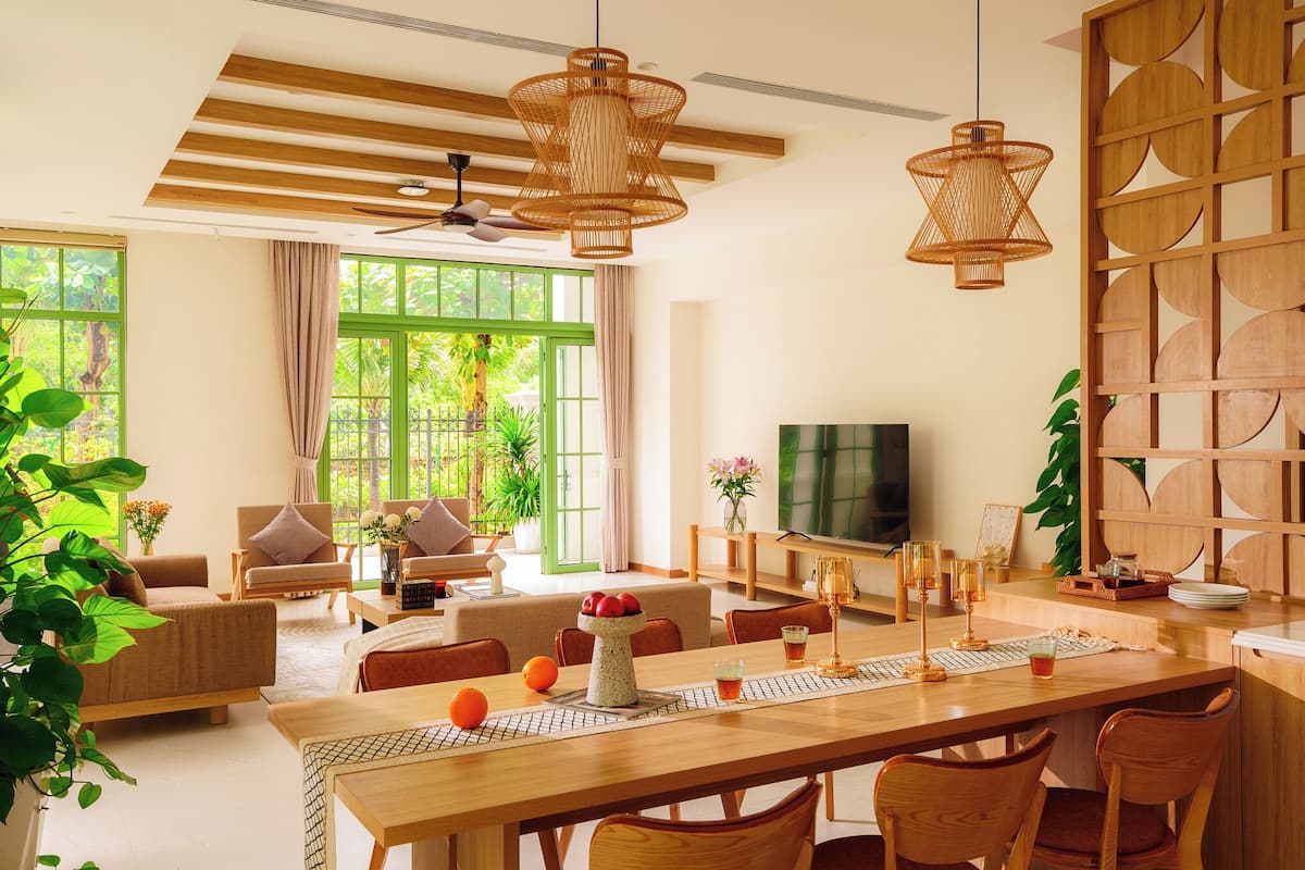 A spacious living room with a wooden dining table, comfortable seating, a television, and large windows with a view of greenery.
