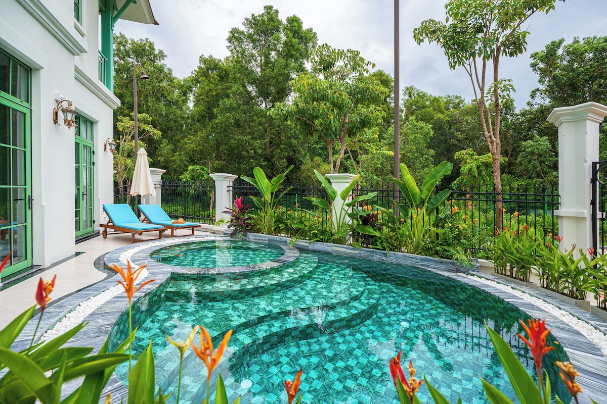 A swimming pool with a tiled design, surrounded by lush greenery and a white building with green accents.