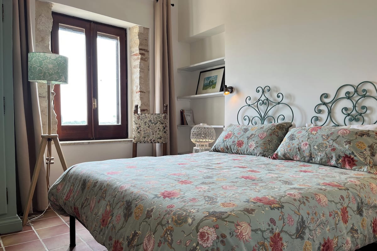A bedroom with a floral bedspread, a standing lamp, a chair, and a metal bed frame.