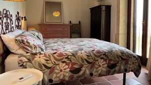 Un dormitorio con colcha estampada, una mesita de noche con control remoto, un aparador, un espejo y un jarrón decorativo.