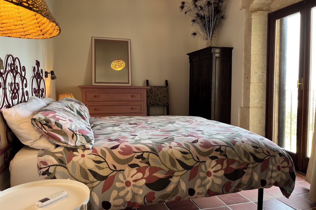 A bedroom with a floral bedspread, a bedside table with a remote control, a dresser, a mirror, and a decorative vase.