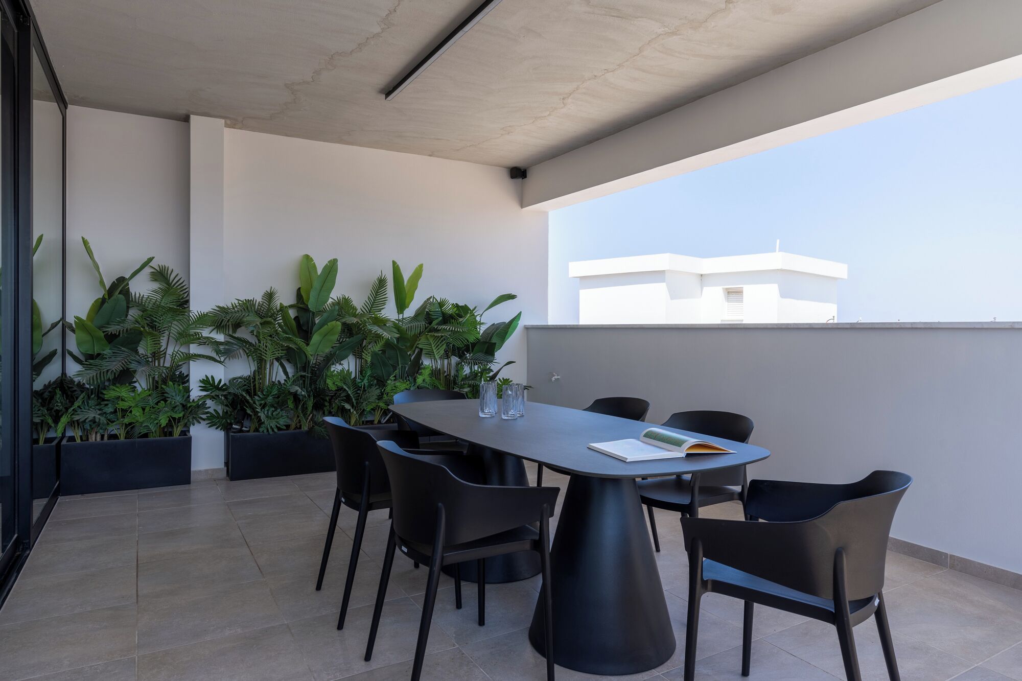 A modern outdoor dining area with a round table and six chairs, surrounded by large potted plants.