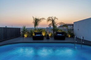 Zona de piscina en la azotea con dos sillones reclinables negros, una mesita con vasos y vistas a un paisaje urbano al atardecer.